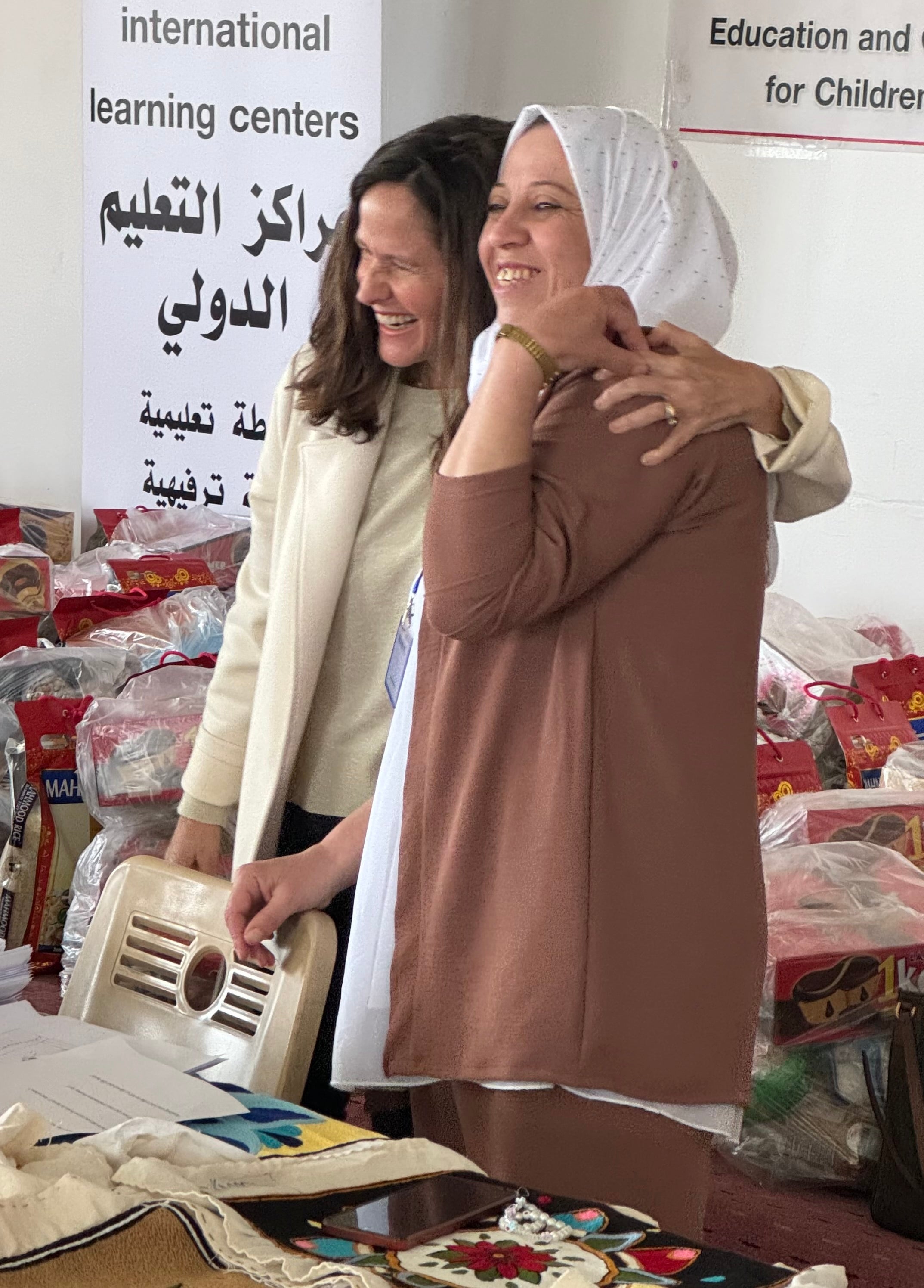 Two women hugging in a room with bags and supplies, with signs in the background.